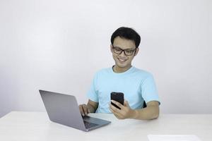 Asian young man happy and smile when he see on smart phone with laptop beside it. Indonesian man wearing blue shirt. photo