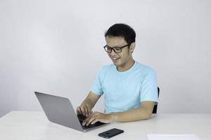 Young Asian man feeling happy and smile when work laptop on table. Indonesian man wearing blue shirt. photo