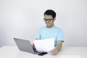 el joven asiático sonríe y es feliz cuando trabaja en una computadora portátil y tiene un documento a mano. hombre indonesio con camisa azul. foto