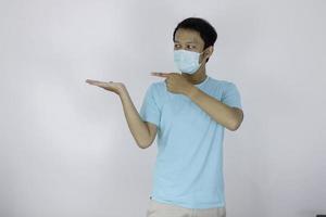 Happy face of young Asian man wear medical mask and blue with hand point on empty space. photo