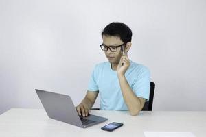 el joven asiático es serio y se enfoca cuando trabaja en una computadora portátil en la mesa. hombre indonesio con camisa azul. foto