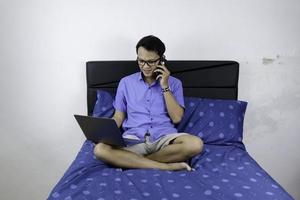 Young asian man is smile and enjoy when working at home with laptop and phone. A man sitting on bed and working. Work from home concept. photo