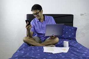 Young asian man is smile and enjoy when working at home with laptop and phone. A man sitting on bed and working. Work from home concept. photo