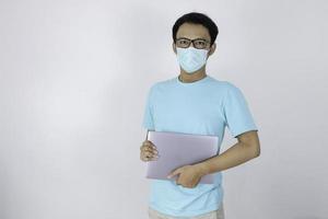 Young Asian Man wear medical mask is carrying a laptop. Indonesian man wearing blue shirt. photo