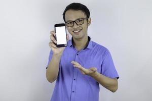 un joven asiático con camisa azul está de pie y sonriendo señalando un espacio en blanco en la pantalla del teléfono inteligente con fondo blanco. foto