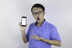 un joven asiático con camisa azul está de pie y se sorprende al señalar un espacio en blanco en la pantalla del teléfono inteligente con fondo blanco. foto