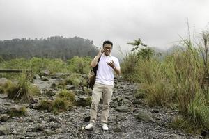 el joven mochilero asiático es feliz y sonríe cuando mira un teléfono inteligente en un viaje de montaña foto