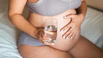 close up of pregnant woman sitting in bed at home holding pills and glass of water. Taking vitamins during pregnancy photo