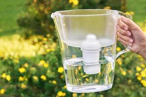 Girl's hand holds a water filter jug in the sunny summer garden with yellow flowers on the background photo