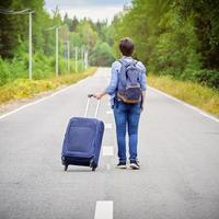 Woman tourist with a travel bag on wheels and a backpack walks away along the narrow road photo