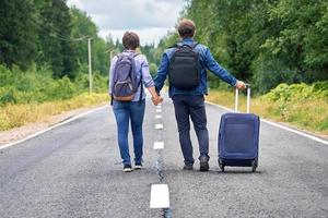 Couple tourists with a travel bag on wheels walk away along the narrow road photo