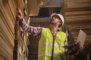 trabajador masculino asiático con uniforme de seguridad y casco, inspector supervisor con computadora portátil para empaquetar pedidos de existencias en el almacén de la fábrica, montones de fabricación de papel apilado, industria de producción de reciclaje. foto