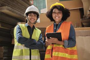 Two female workers and colleagues in safety uniforms and helmet stock check, control production in storage of warehouse factory with a lot of paper stack, friend works in recycle manufacture business. photo