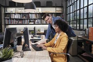 Young African American female worker sits, works with computer, brainstorming, talking, and discussing with Caucasian male colleague and partnership about business jobs in workspace office company. photo