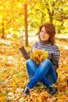 hermosa mujer alegre de moda sentada en hojas amarillas en el parque de otoño y sosteniendo las hojas en sus manos foto