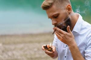 retrato de un apuesto hombre barbudo, enciende un cigarrillo con un encendedor al aire libre foto