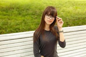 retrato de una linda chica con gafas, sentada en un banco en la calle en el parque foto