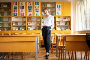 una hermosa chica con una camisa blanca se para en el salón de clases entre los escritorios foto