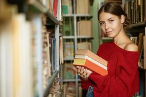 mujer joven sosteniendo una pila de libros en el fondo de la biblioteca. foto