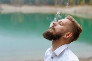 hombre de negocios caucásico joven barbudo en una camisa blanca fuma un cigarrillo foto
