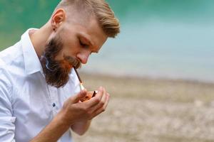 retrato de un apuesto hombre barbudo, enciende un cigarrillo con un encendedor al aire libre foto