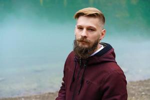 Young bearded man stood alone by lake and looked aside thoughtfully smoking photo