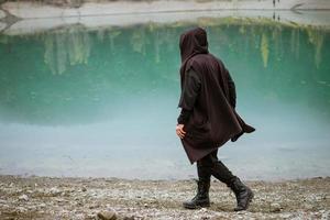 A man in a sweatshirt with a hood walks alone on the beach by a calm lake photo