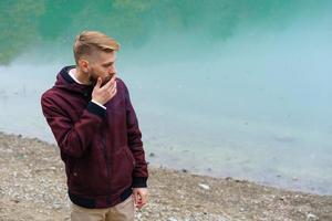 un joven barbudo estaba solo junto al lago y miraba a un lado fumando pensativamente foto