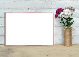 Rose Gold horizontal A4 Frame mockup near a bouquet of sweet-william stands on a wooden table on a painted white wooden background. Rustic style, simple beauty. 3d render. photo