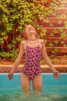 la niña se para en la piscina y disfruta del clima soleado de verano. adolescente en traje de baño. foto