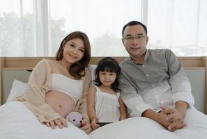 retrato de una familia embarazada, incluida la madre, el padre y la hija, sentados juntos en la cama