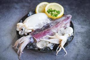 calamares crudos con hielo y especias de ensalada limón en el fondo del plato negro - calamares frescos pulpo o sepia para comida cocinada en el restaurante o mercado de mariscos foto