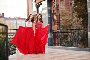 Portrait of two fashionable girls at red evening dress posed background mirror window of modern building photo