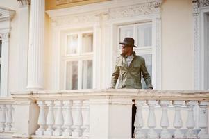 retrato de moda de hombre afroamericano negro con capa de abrigo verde y sombrero negro, quédese en el balcón de la mansión amarilla foto