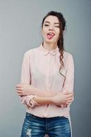 Portrait of young brunette girl showing tongue and winks, wearing in pink blouse, ripped jeans. Fashion studio shot