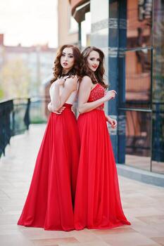 Portrait of two fashionable girls at red evening dress posed background mirror window of modern building photo