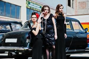 tres muchachas vestidas de estilo retro cerca de viejos autos antiguos clásicos. foto