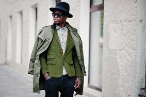 Fashion portrait of black african american man on green velvet jacket, black hat and coat cloak on his shoulders walking on streets of city background house with many windows photo