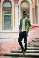 Fashion portrait of black african american man on green velvet jacket and black hat stay on stairs background old mansion. Vertical photo