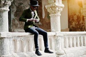 Fashion portrait of black african american man on green velvet jacket and black hat, sitting on the railing background old vintage house with arch photo