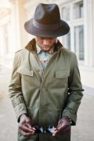 Fashion portrait of black african american man on green coat cloak and black hat, stay on the balcony of yellow mansion. Vertical photo