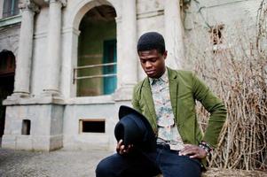 Fashion portrait of black african american man on green velvet jacket and black hat, sitting at felled tree background old vintage house with arch and columns photo