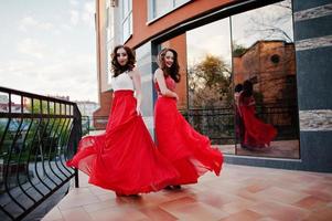 Portrait of two fashionable girls at red evening dress posed background mirror window of modern building photo