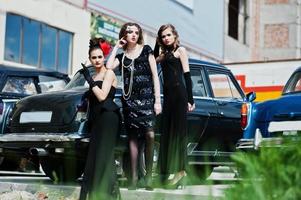 tres muchachas vestidas de estilo retro cerca de viejos autos antiguos clásicos. foto
