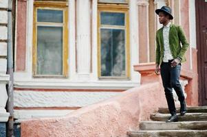 Fashion portrait of black african american man on green velvet jacket and black hat stay on stairs background old mansion. photo