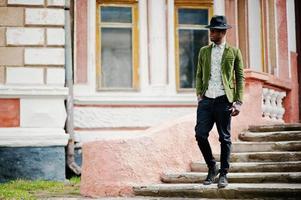 Fashion portrait of black african american man on green velvet jacket and black hat stay on stairs background old mansion. photo