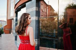 Portrait back of fashionable girl at red evening dress posed background mirror window of modern building photo