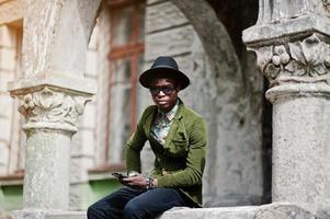 Fashion portrait of black african american man on green velvet jacket and black hat, sitting on the railing and look at camera, holding his smartphone at hand background old vintage house with arch photo