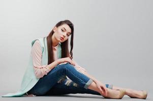 Sitting portrait young brunette girl wearing in pink blouse, turquoise jacket, ripped jeans and cream shoes .Fashion studio shot on gray background photo
