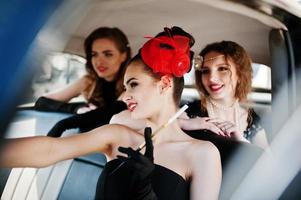 Three young girl in retro style dress seat on old classic vintage car and having fun emotions photo